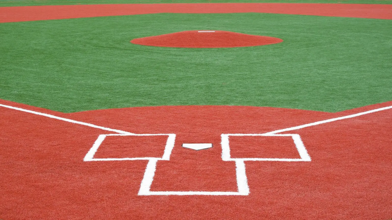 A close-up view of a baseball field shows the home plate, batter's boxes, pitcher's mound, and surrounding green and red artificial turf by Goodyear Greens and Turf. The vibrant colors highlight the infield's distinct boundaries and features.