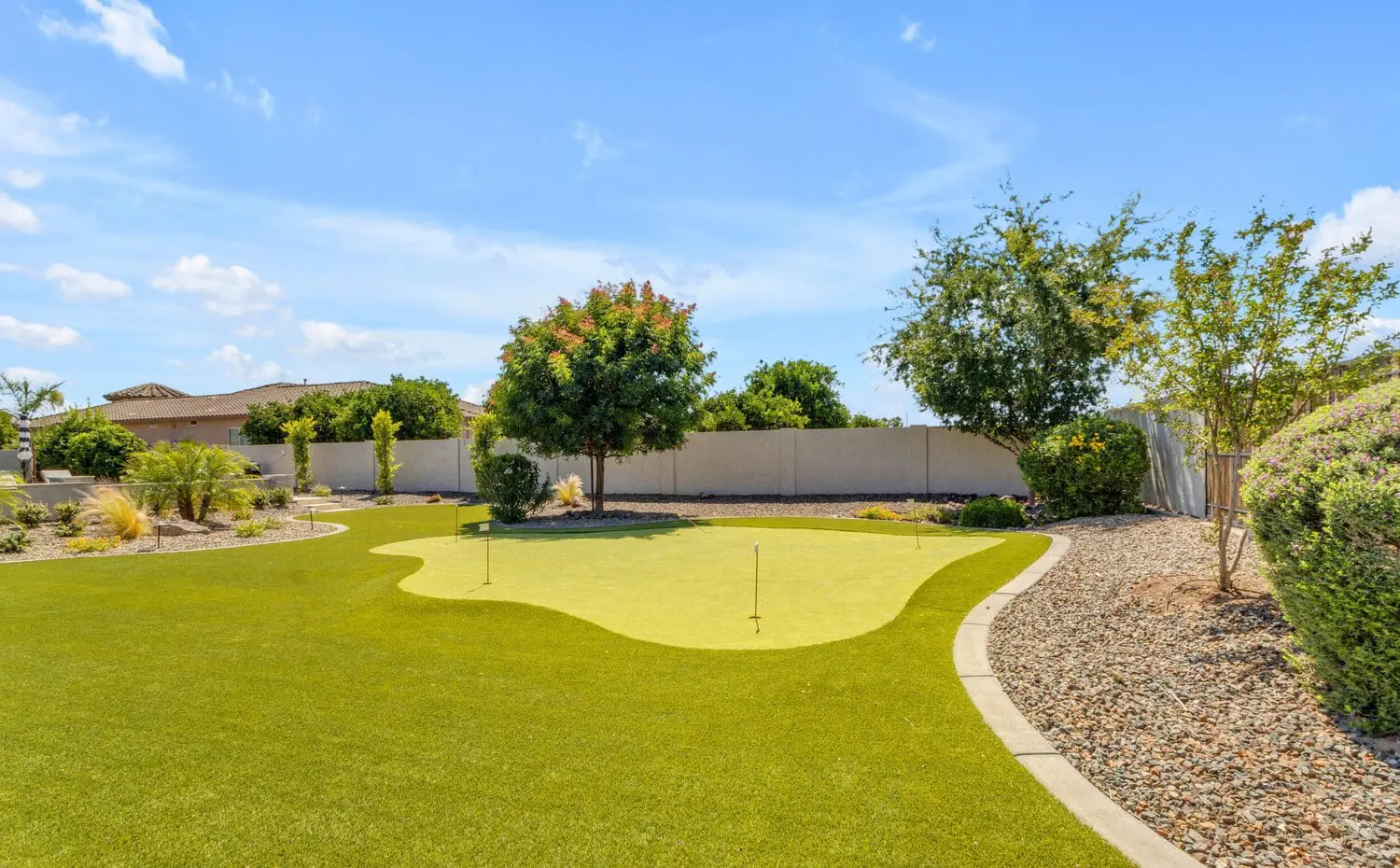 A well-maintained backyard in Goodyear, AZ, featuring a putting green with athletic turf installer precision, surrounded by neatly trimmed hedges and trees. A rock garden borders the green, and a beige wall frames the background with a house partially visible on the left. The sky is clear and blue.