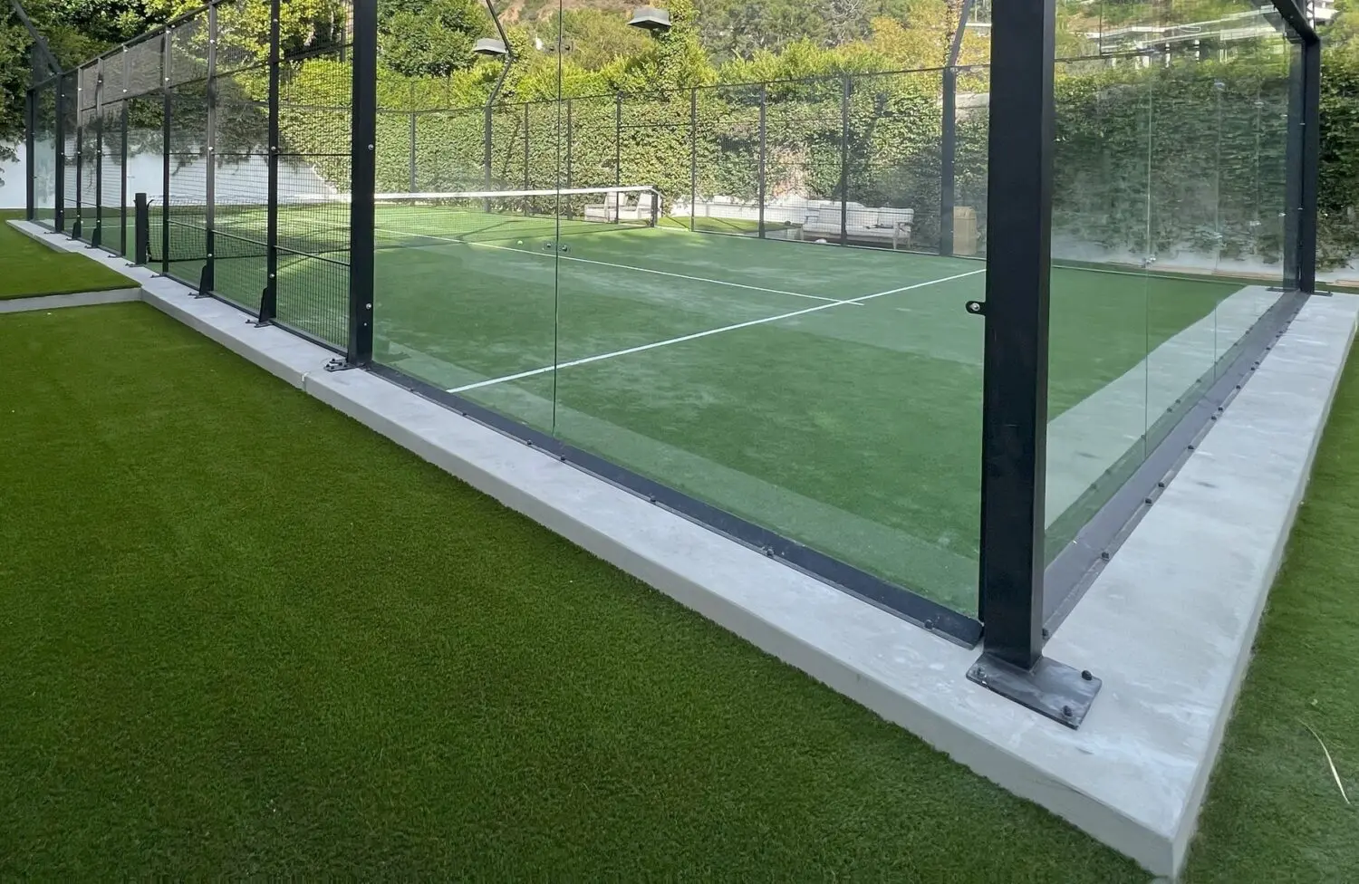 An outdoor padel court with a glass enclosure on a sunny day. Featuring Goodyear Greens and Turf's well-maintained green artificial turf surface, the court is surrounded by lush green hedges and trees. In the background, a bench and a net are visible inside the court.