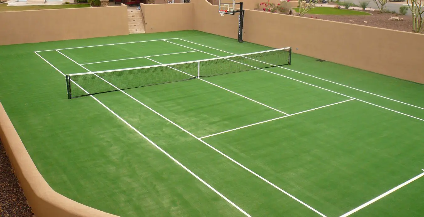 An outdoor sports court with multi-purpose lines for tennis and possibly other sports features a tennis net in the center and a basketball hoop mounted on one of the beige walls. The court, installed by a skilled athletic turf installer, is enclosed by desert landscaping.