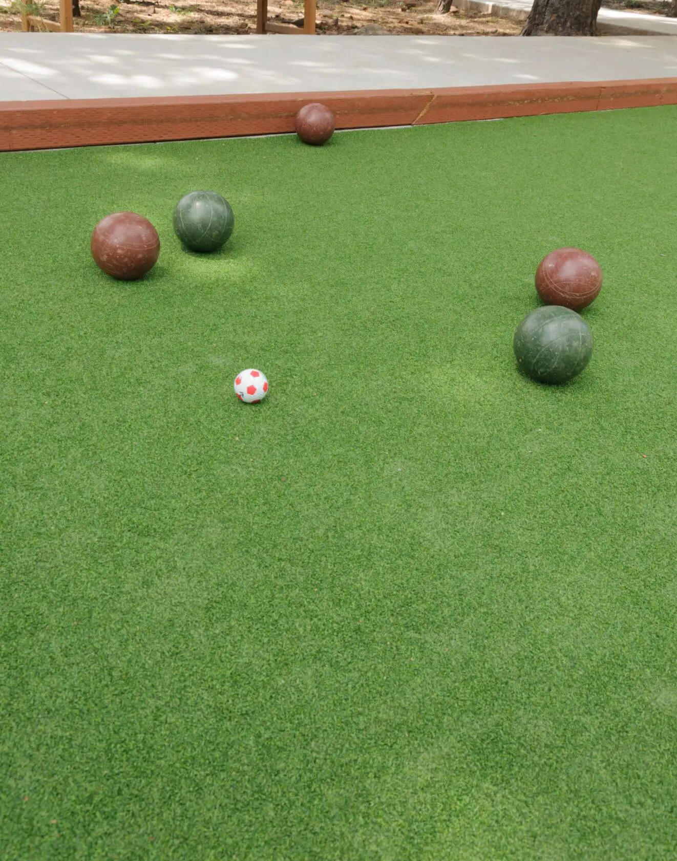 A bocce ball court with six balls on green turf. There are two brown balls, two green balls, and one white target ball with red spots scattered on the field. An athletic turf installer from Goodyear AZ has expertly laid the sports field artificial turf bordered by a wooden boundary at the back.