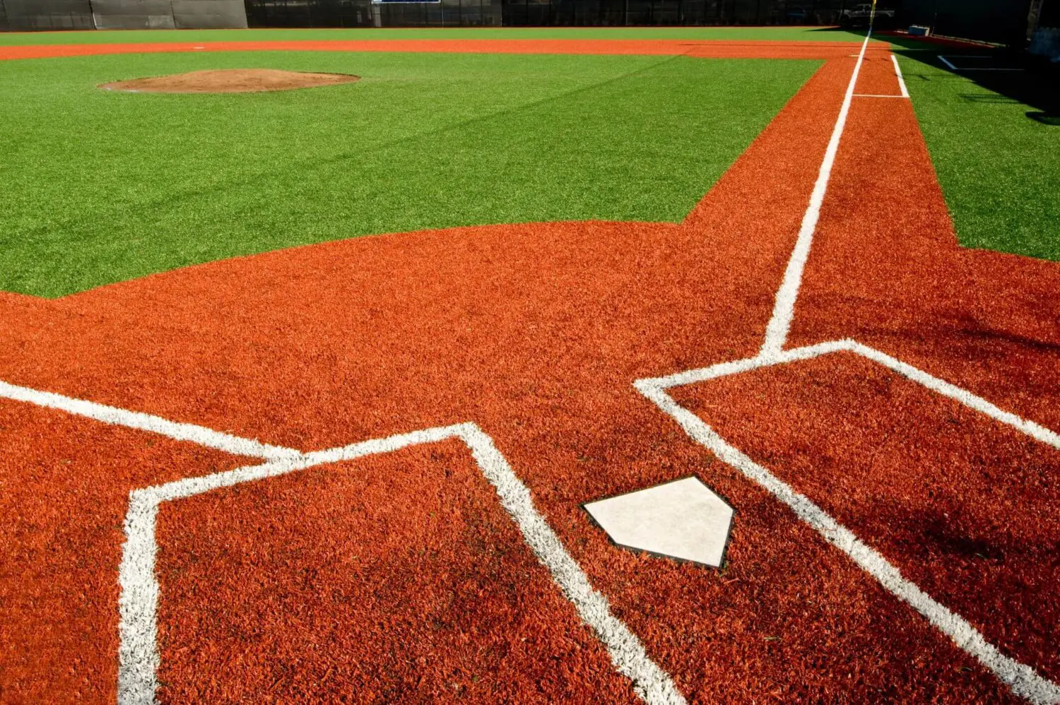 A close-up view of a baseball field featuring home plate, the batter's boxes, and the pitcher's mound. The field boasts red artificial turf around the home plate area from Goodyear Greens and Turf, which contrasts with the green turf of the rest of the field. White lines mark the boundaries.