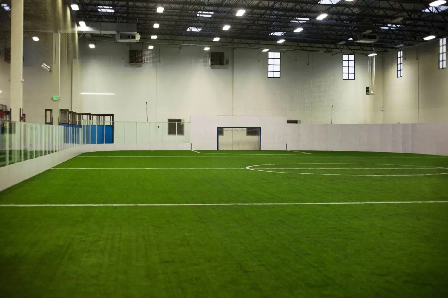 An indoor soccer field with professional-grade artificial turf, surrounded by white walls and glass barriers. The ceiling has exposed beams and several lights. Windows on the walls allow natural light to enter. The goalpost is visible, and the renovated sports field is well lit.