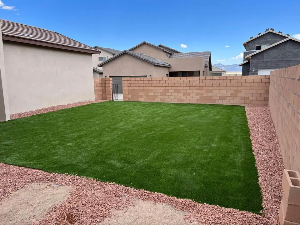 A backyard with a neatly maintained lawn, enhanced by an expert artificial grass installation, surrounded by a border of red gravel and enclosed by a tan cinder block fence. The sky is clear with a few clouds, and neighboring houses are visible in the background.