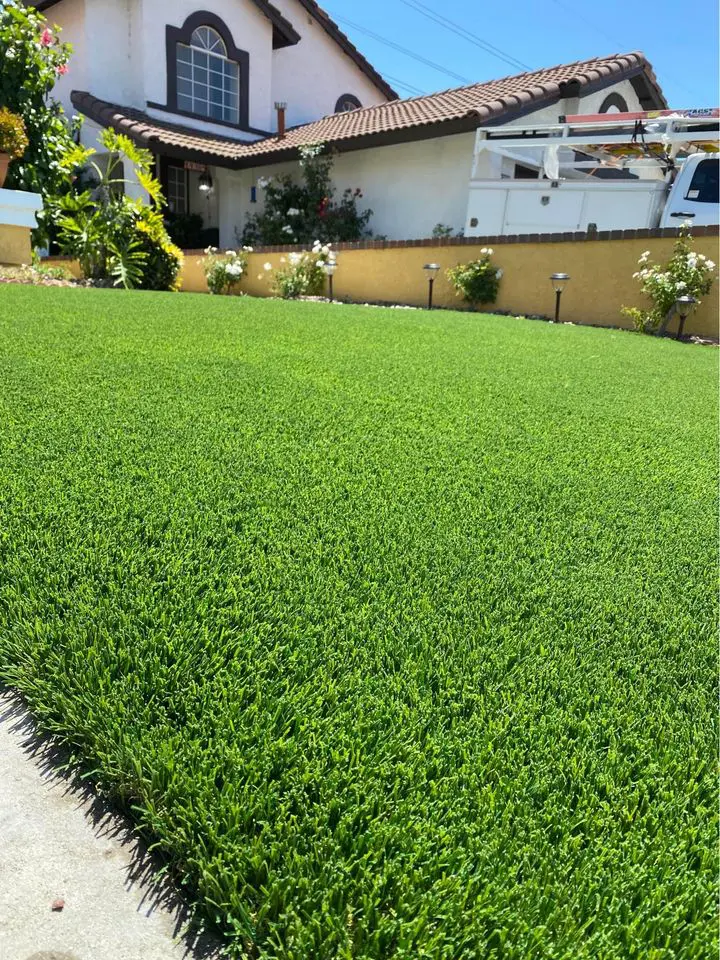 Close-up of a narrow, neatly trimmed green lawn bordered by a low stone retaining wall on the right and the side of a white building on the left. The wall and building create a corridor effect, showcasing artificial grass by Scottsdale Turf Pros under a clear blue sky.