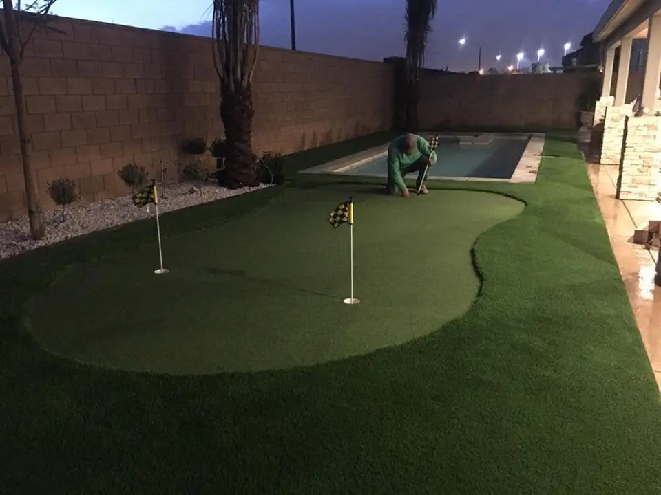 A person kneels on a Goodyear Greens Turf backyard putting green, adjusting a flag. The area includes two holes with checkered flags. A narrow swimming pool is to the right, and the scene is illuminated in the evening light, showcasing a flawless artificial grass installation.