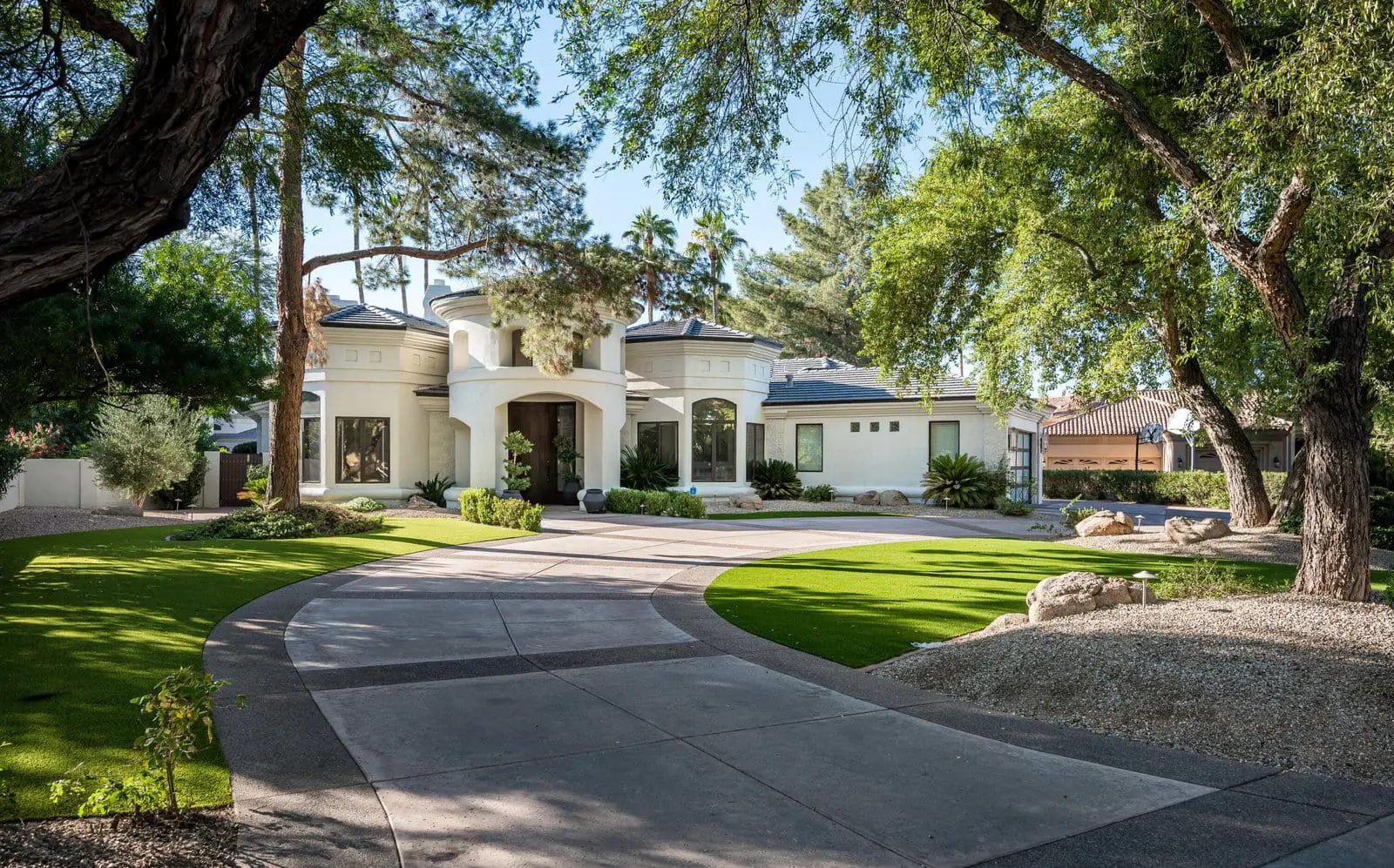 A modern, two-story white house with large windows, nestled amid lush green trees and landscaping. A wide, curved driveway leads to the entrance, surrounded by a manicured lawn featuring synthetic grass and well-maintained foliage. The scene appears serene and inviting.