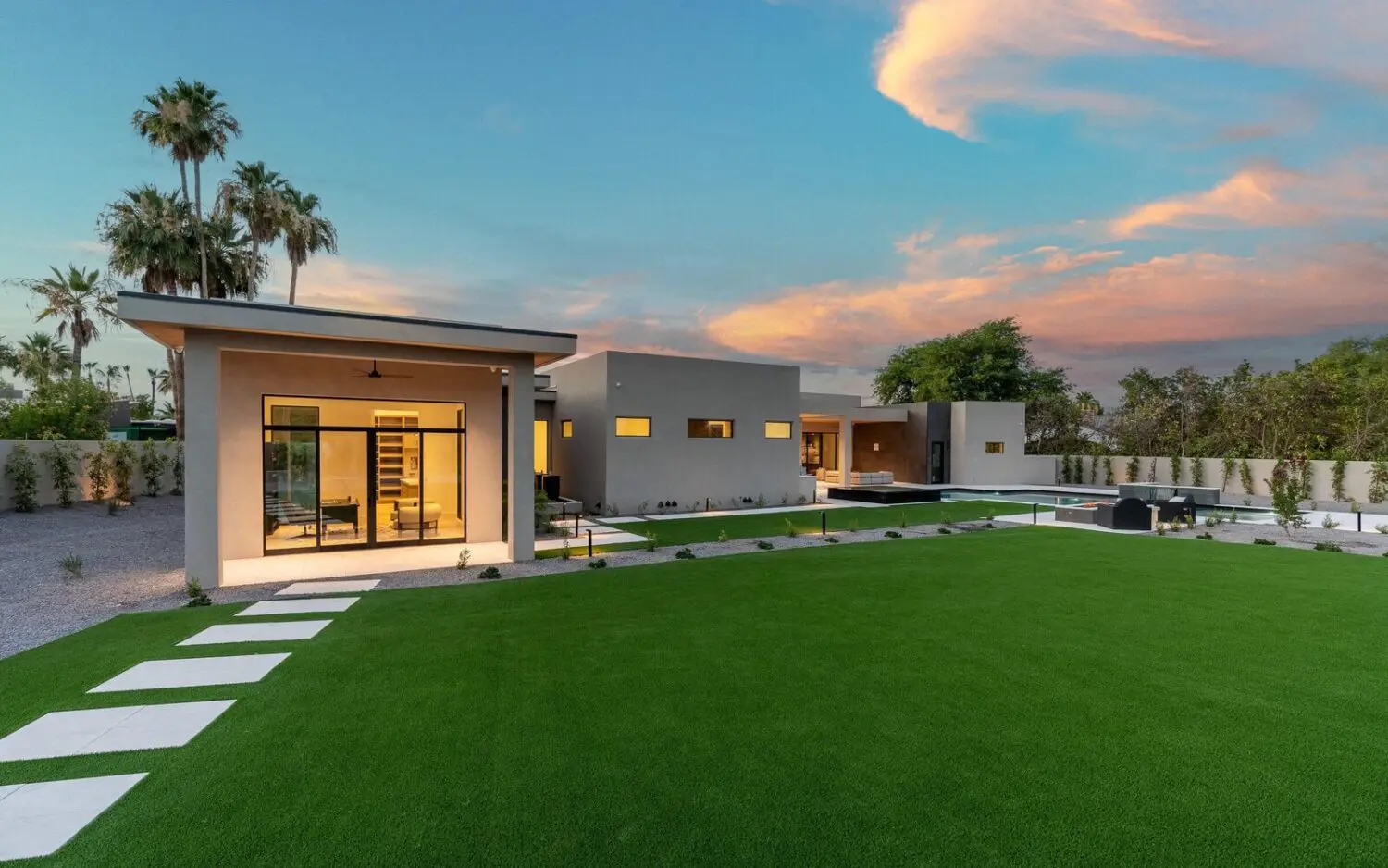 A modern, single-story house with large glass windows and a flat roof sits in a well-manicured yard featuring synthetic grass and palm trees. A paved pathway leads to the entrance. The sky is partially cloudy with a warm, colorful sunset. White stone pathways cut through the Goodyear landscape.