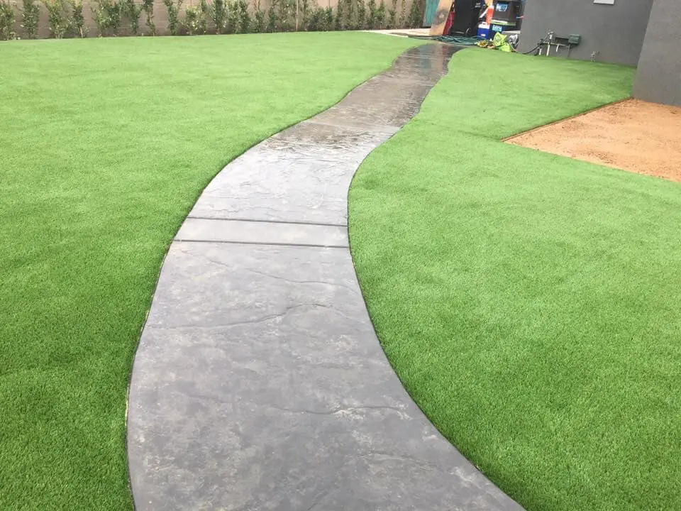 A winding stone path runs through a well-manicured lawn with bright green synthetic grass from Goodyear Greens Turf. The path appears shiny as if wet from recent rain. In the background, there is a garden bed and some garden tools near a building.