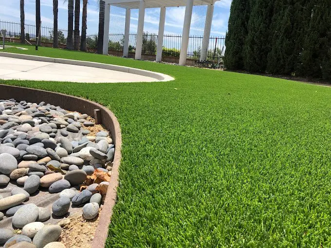 A well-maintained front yard with artificial grass, featuring a dry riverbed made of pebbles and bordered by large rocks. The area includes a tree, trimmed bushes, and a driveway leading to a garage with an old Volkswagen Beetle. Scottsdale Turf Pros completed the pet turf installation. Another house is visible in the background.