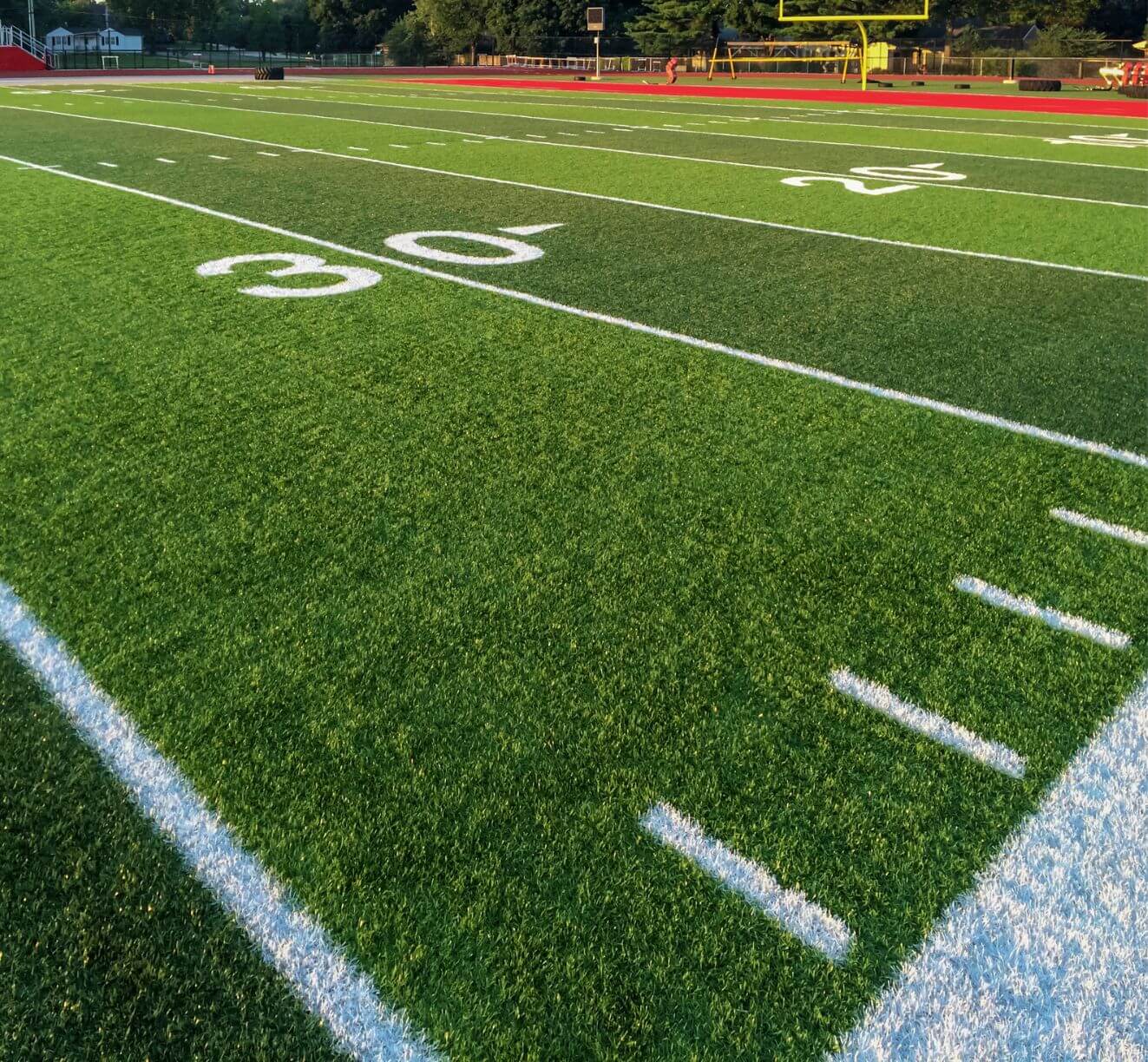 A close-up view of a football field showing the 30-yard line, white yard markers, and a portion of the end zone. The field is marked with bright white lines and green turf by Goodyear Greens and Turf, with goalposts and stadium seating visible in the background.